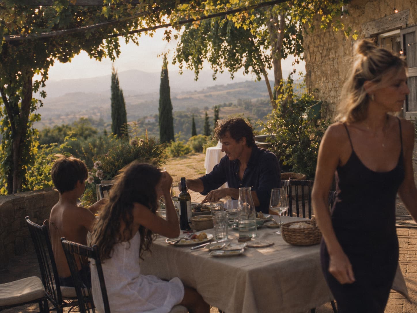 A family sharing a meal beneath a vine-covered pergola overlooking the Tuscan hills.